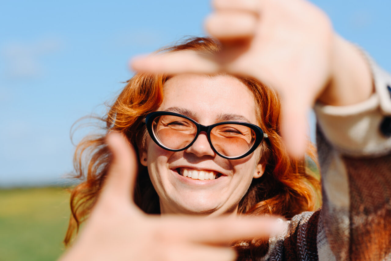 woman standing on country road, shows camera gesture with fingers, beige plaid shirt, sunglasses, sunny summer day, blue sky, green field and trees, smiling happily